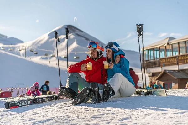 Ein Paar in Skibekleidung sitzt im Schnee und trinkt heiße Getränke vor einer Berghütte.